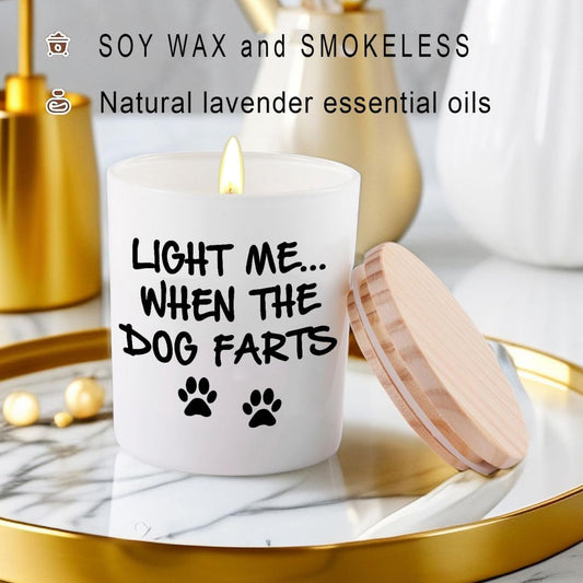 A white candle with a lit flame sits on a marble tray with a wooden lid beside it. The candle has black text reading 'Light me when the dog farts.'