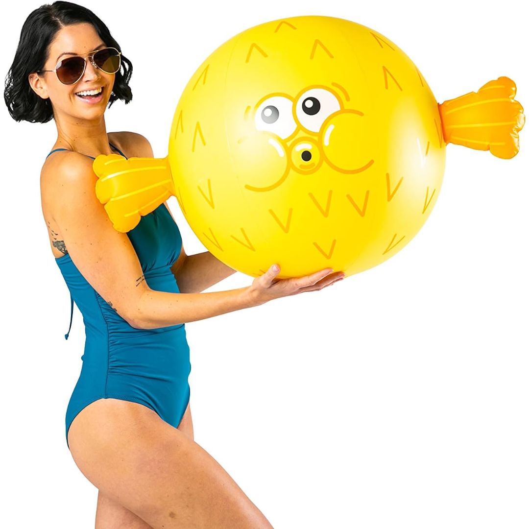A smiling woman with sunglasses holding a bright yellow inflatable beach ball shaped like a puffer fish 