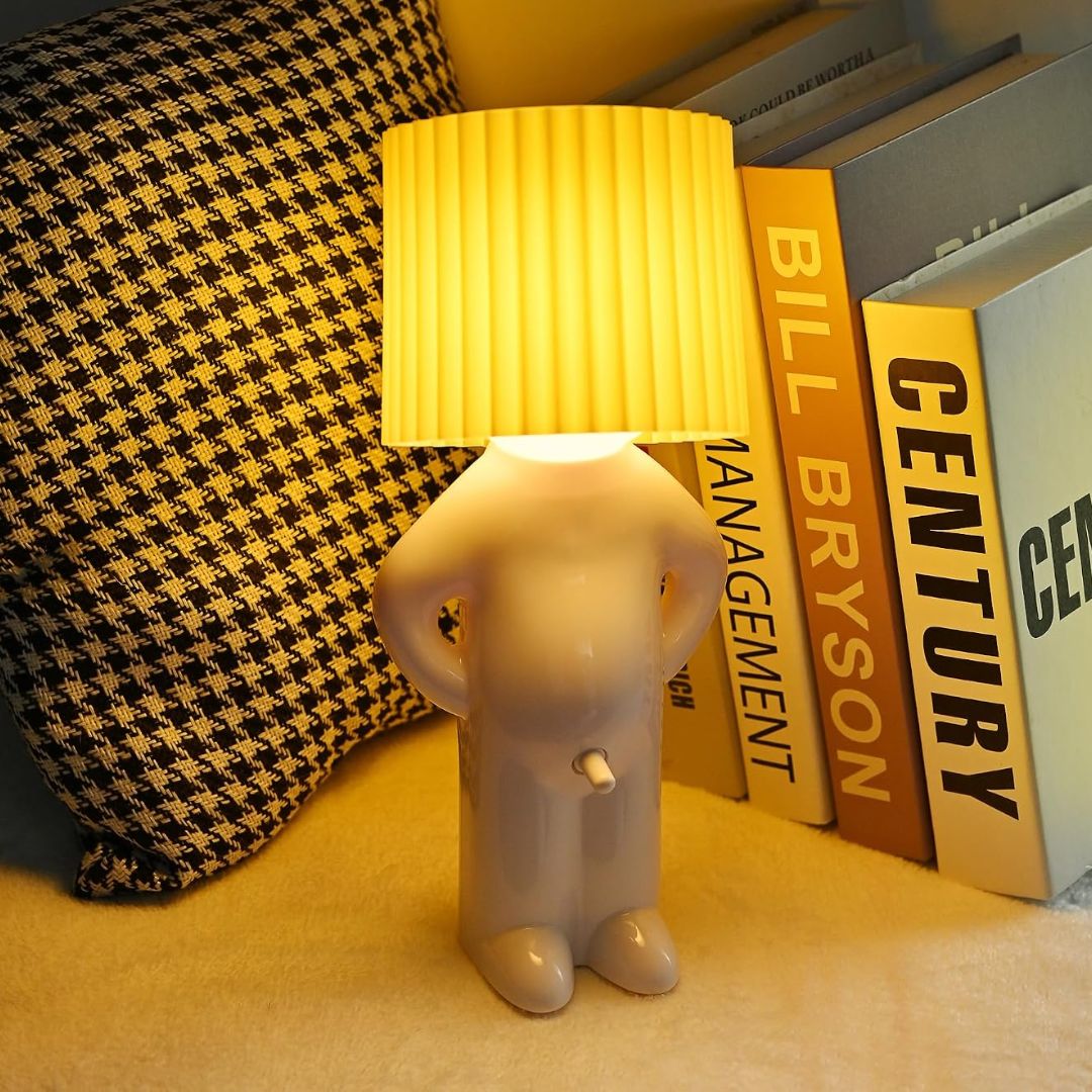 A novelty table lamp shaped like a small human figure with a pleated yellow lampshade for a head, glowing on a tabletop beside books and a houndstooth pillow.