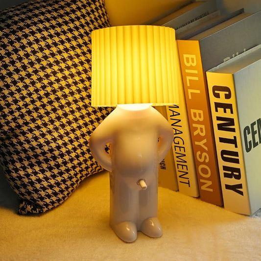 A novelty table lamp shaped like a small human figure with a pleated yellow lampshade for a head, glowing on a tabletop beside books and a houndstooth pillow.