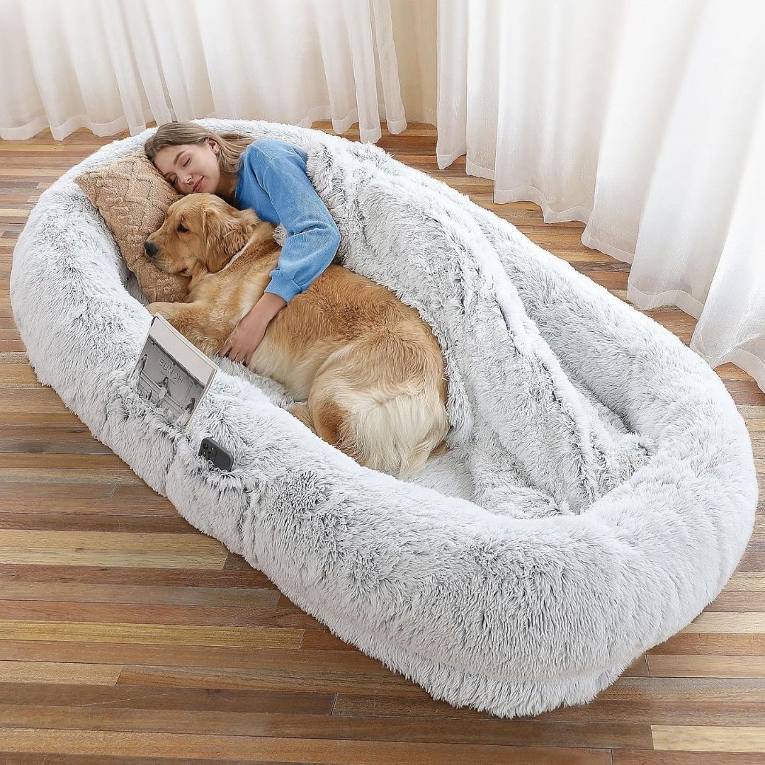 Woman hugging a dog in a fluffy human pet bed on a wooden floor.