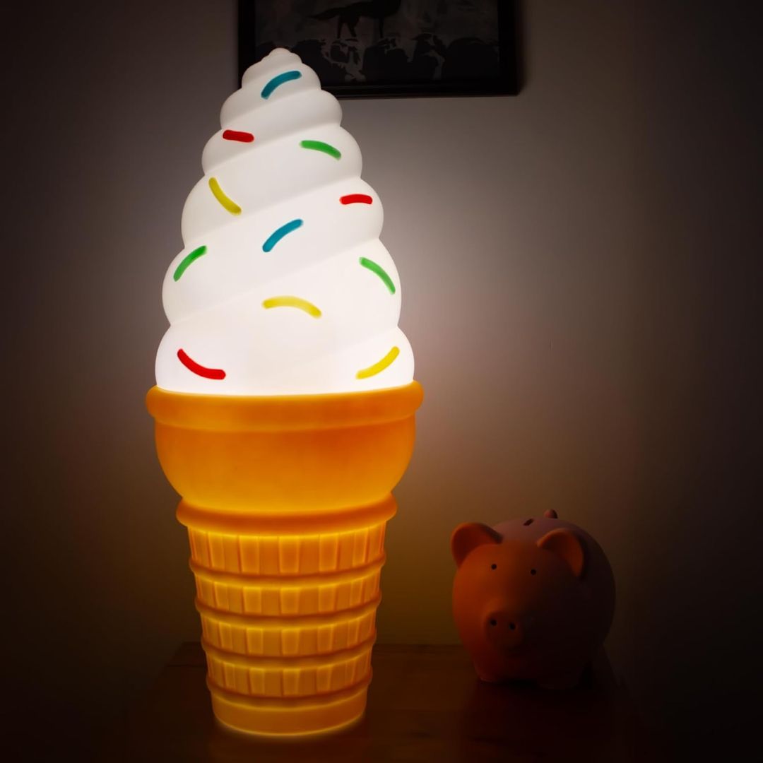 A large novelty lamp shaped like a soft-ice cream cone, glowing with a warm yellow-orange waffle-cone base and a white swirled top decorated with small multicolored candy-like sprinkles, shown on a dark tabletop next to a small pig figurine.