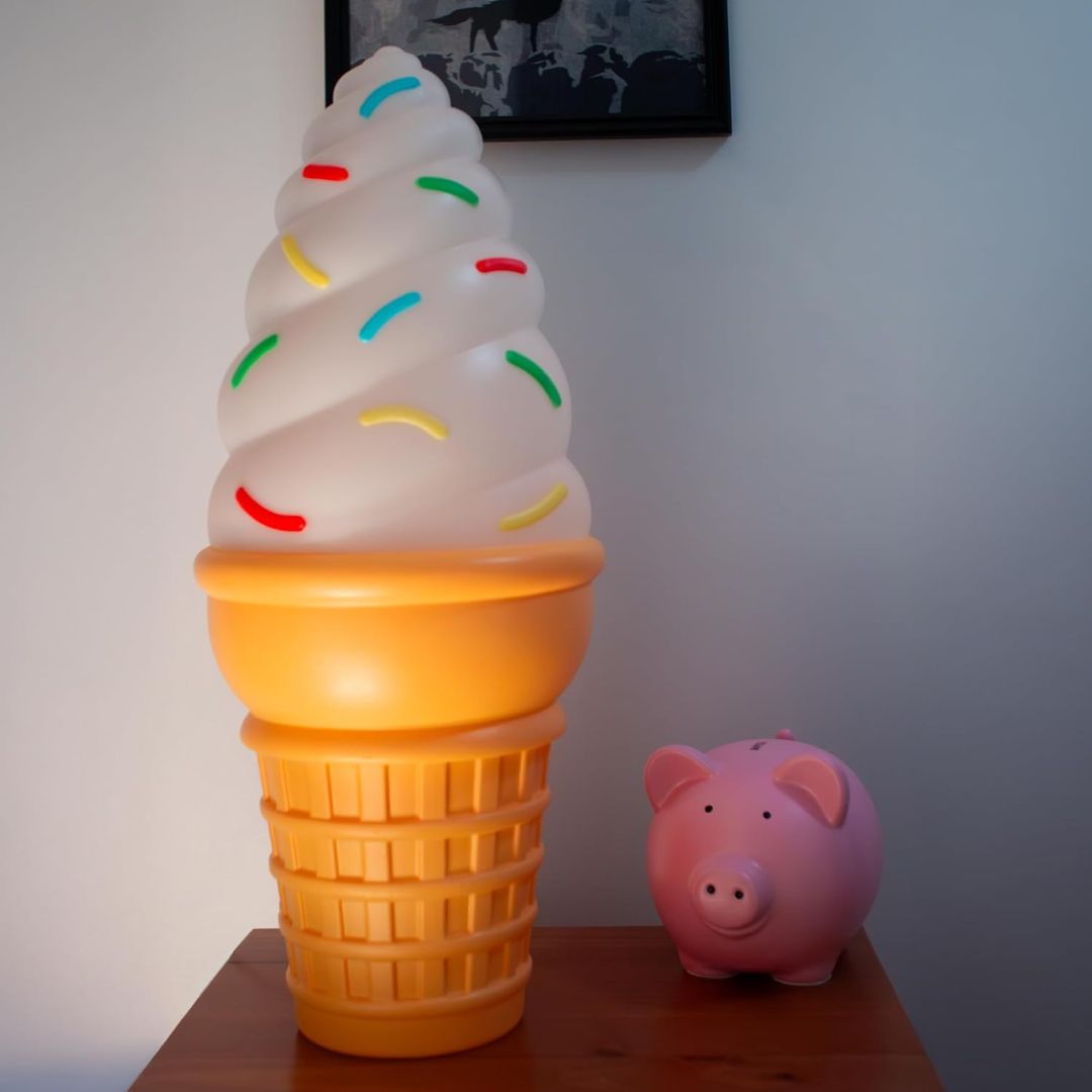 A novelty lamp shaped like a soft-ice cream cone with a glowing yellow-orange cone base and a white swirl top decorated with multicolored sprinkles, shown on a wooden table next to a pink piggy bank against a gray wall.