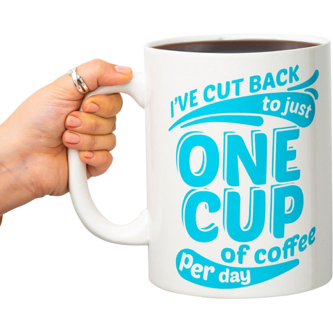 Giant white coffee mug with blue text 'I've cut back to just ONE CUP of coffee per day' held by a hand.