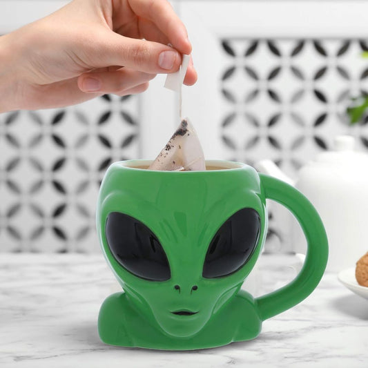 A green alien-head ceramic mug with large black eyes, shown on a marble surface with a hand inserting a tea bag into the cup.