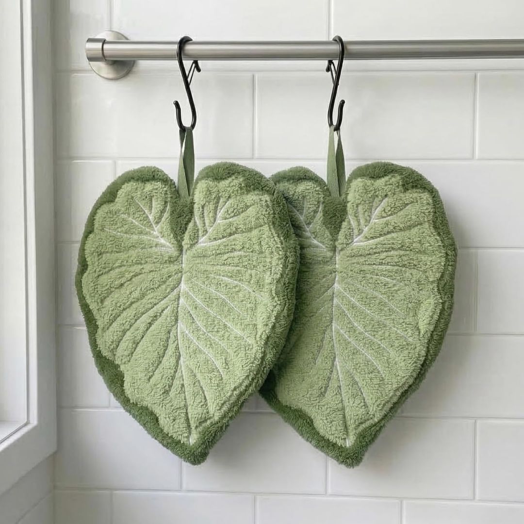 Two green heart-shaped hand towels with leaf-like embossed patterns hang from black hooks on a metal rod against a white tiled wall.