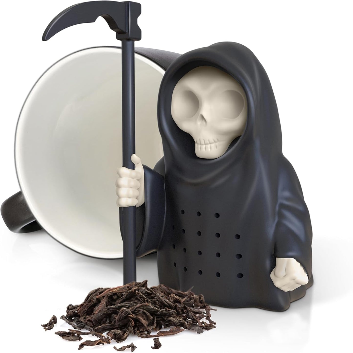 A black silicone Grim Reaper tea infuser with a scythe, next to a white mug and scattered loose tea leaves on a white surface.