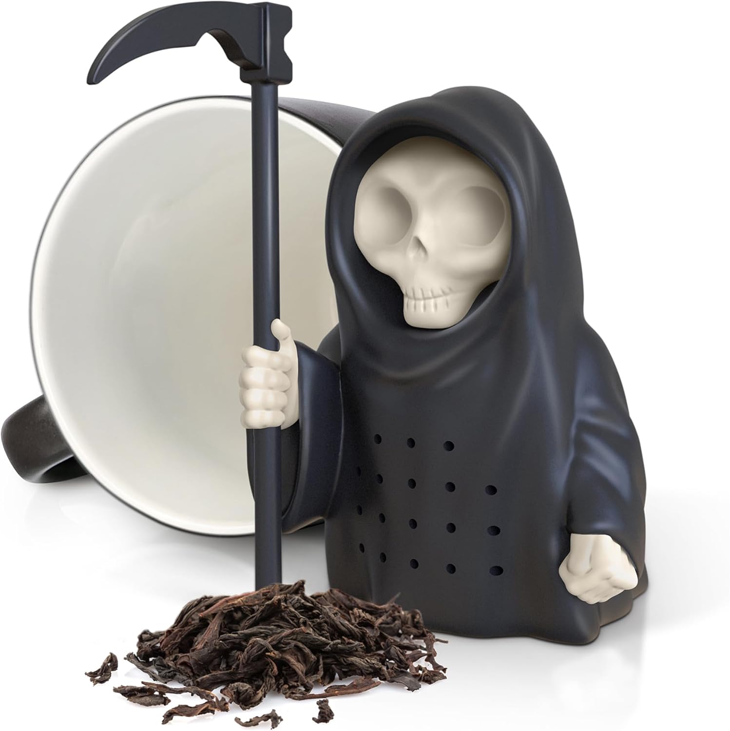 A black silicone Grim Reaper tea infuser with a scythe, next to a white mug and scattered loose tea leaves on a white surface.