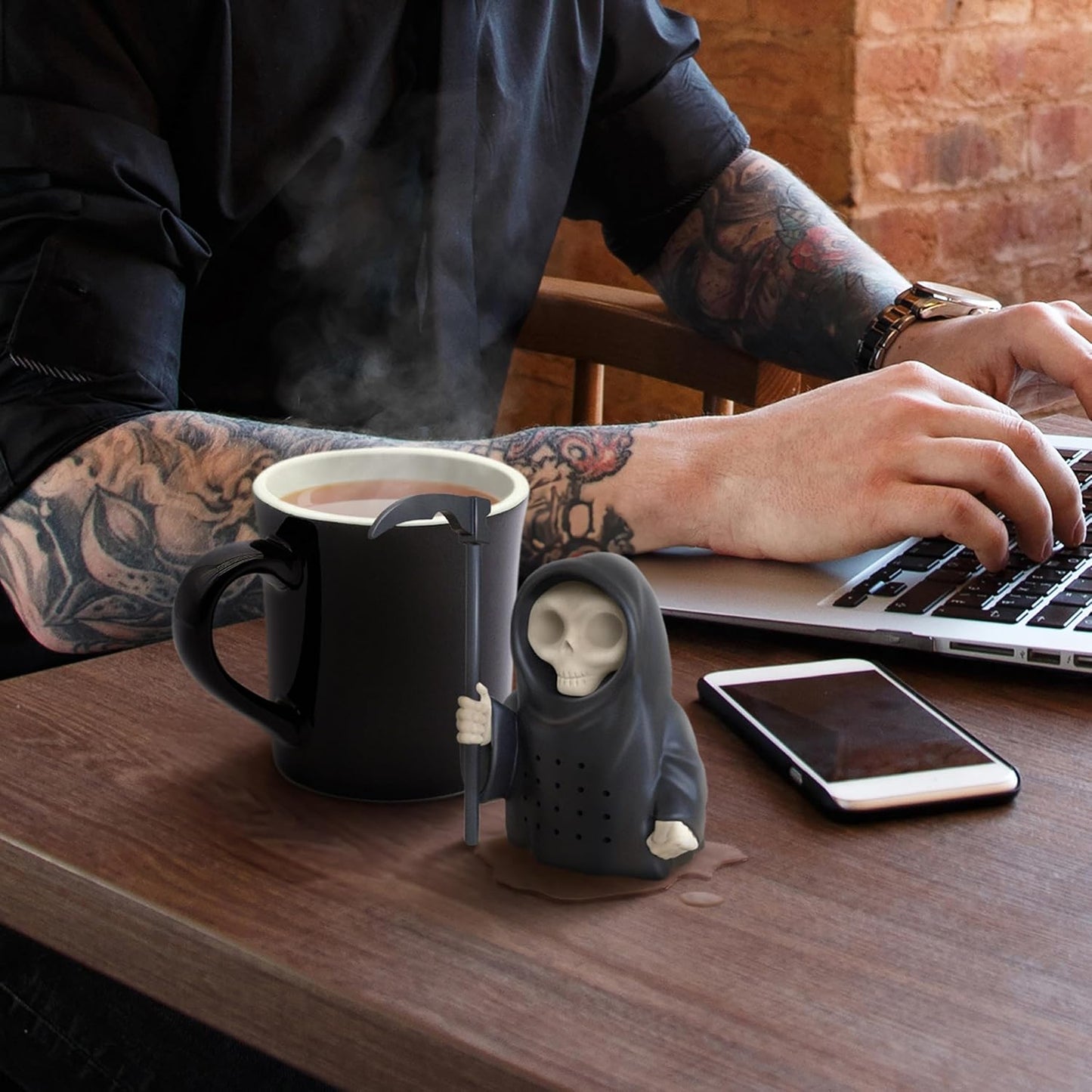 A person with tattooed arms works on a laptop, with a black mug of steaming tea, a Grim Reaper tea infuser, and a smartphone on a wooden table.