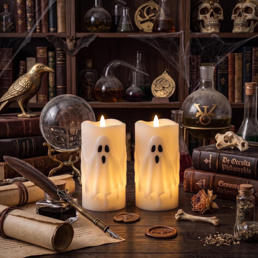 Two glowing cream ghost-shaped flameless pillar candles with black facial features, displayed on a dark wooden table surrounded by occult-themed props including old books, a quill, ink, dried flowers, and a glass orb, with a dark bookshelf in the background.