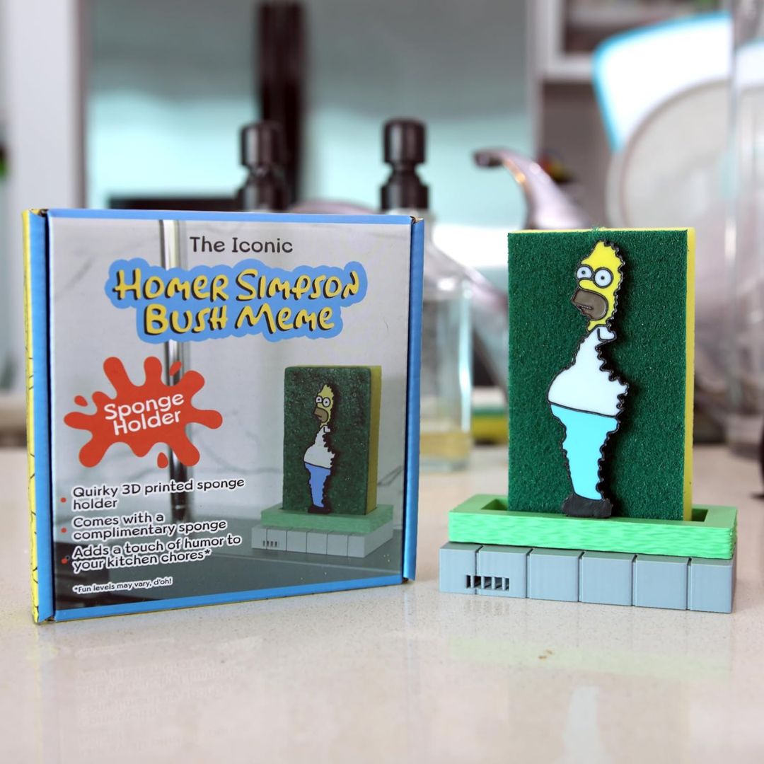 Homer Simpson sponge holder featuring a Homer Simpson character sponge and a green cleaning sponge on a light blue and green pixelated base, shown next to its retail packaging on a kitchen counter.