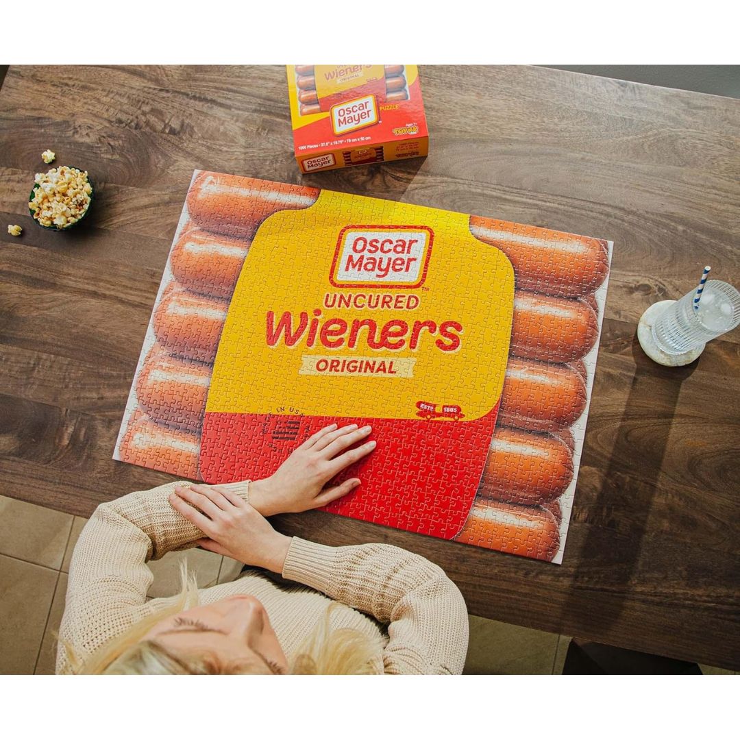 A person sits at a wooden table with their hands on a very large Oscar Mayer Uncured Wieners Original jigsaw puzzle. A real box of Oscar Mayer Wieners is visible in the background along with a bowl of popcorn and a drink.