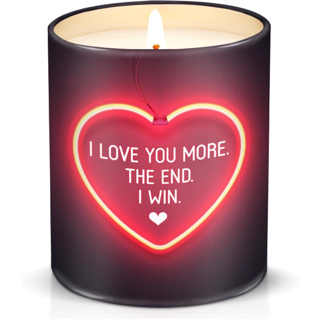 A dark purple cylindrical candle with a lit wick and a glowing red heart-shaped neon design on the front that reads, 'I LOVE YOU MORE. THE END.