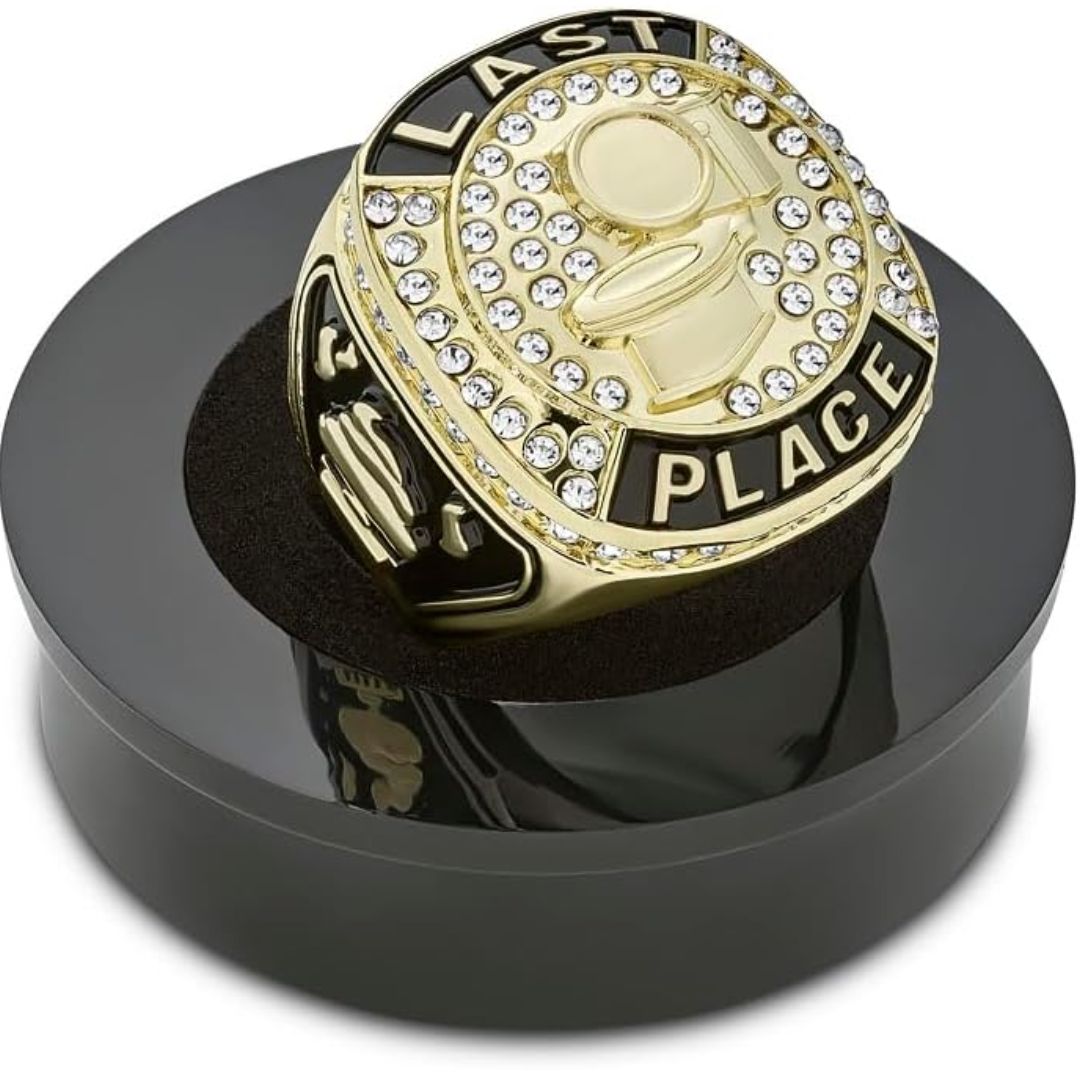 A gold-plated championship ring with black enamel lettering reading 'LAST PLACE' and a toilet icon and surrounded by clear rhinestones, displayed on a black circular stand.