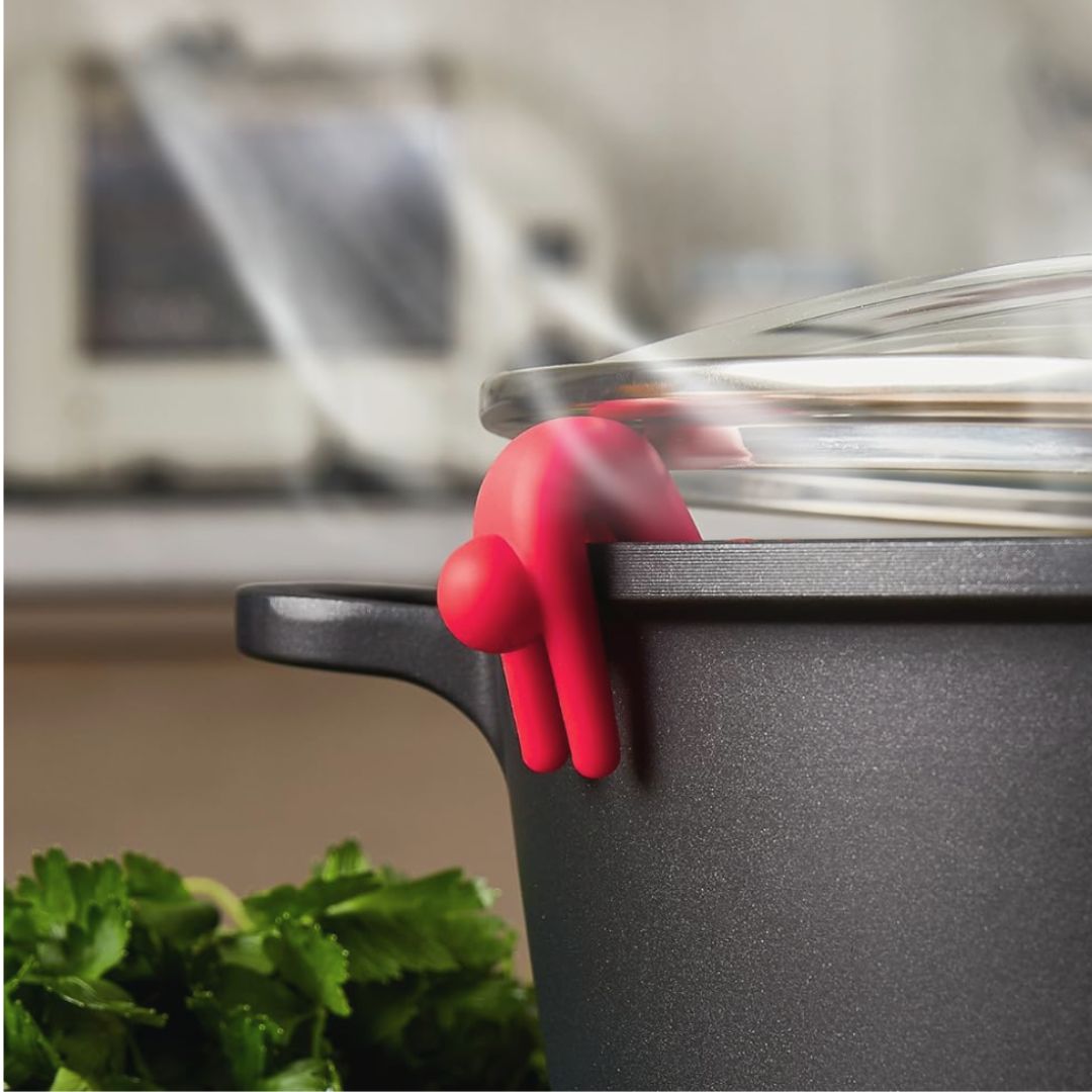 Red silicone pot holder shaped as a slumped over man on a black pot with a blurred kitchen background
