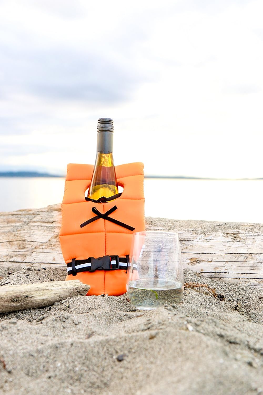 Wine bottle dressed in an orange life jacket cover on a sandy beach with a driftwood log and a glass of white wine nearby.