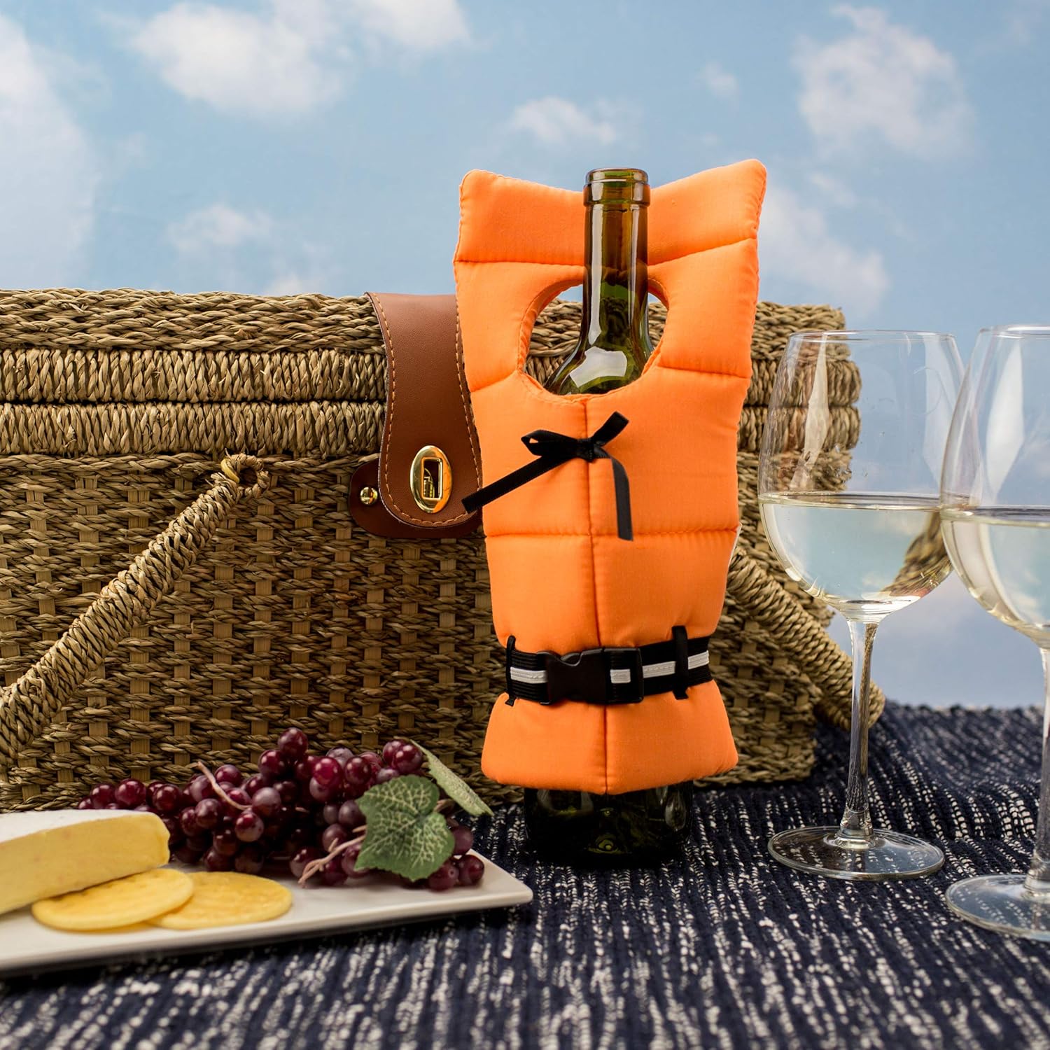 Wine bottle in an orange life jacket cover with picnic basket, cheese, grapes, and two glasses of white wine on a sunny day.