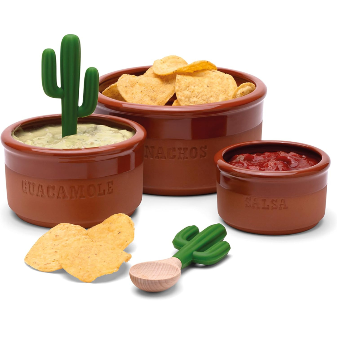 Three terracotta-style bowls labeled guacamole, nachos, and salsa with chips and a cactus-shaped spoon and dip stick.