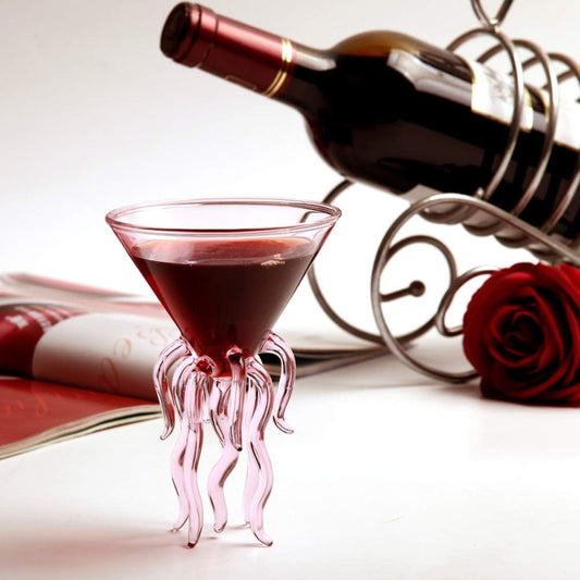 Pink octopus-shaped glass with red liquid next to a bottle of red wine and a rose on a white surface.