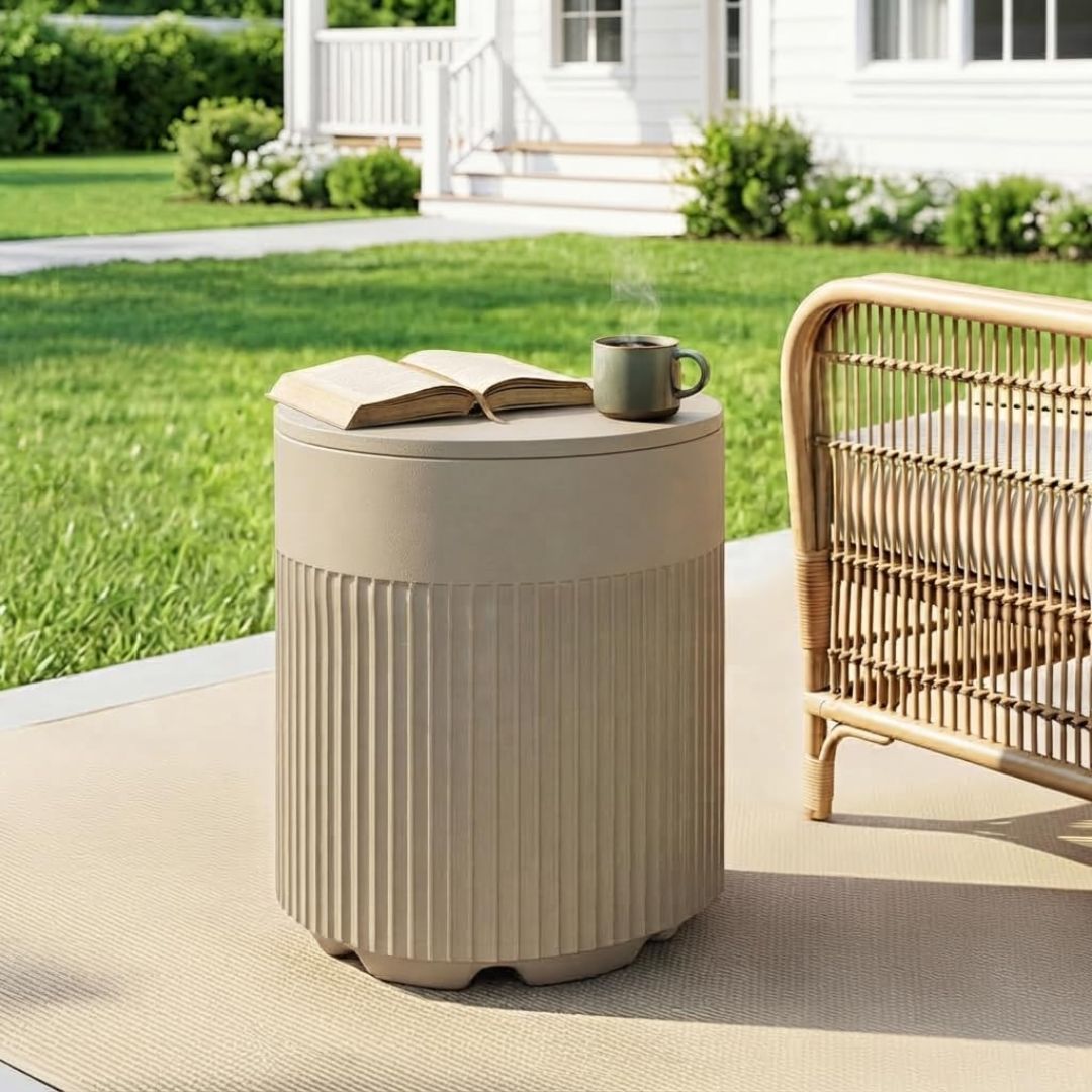 A beige patio umbrella base side table with a ribbed lower section and a hinged lid, shown on a patio with an open book and mug on top. A wicker chair is partially visible beside it, with a lawn and house in the background.