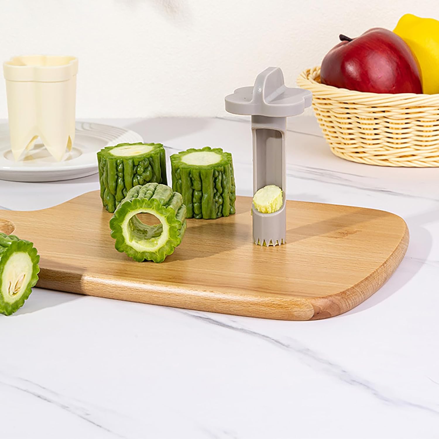 A pepper seed corer remover as it cores green okra on a wooden board, with cored okra pieces, a basket of apples, and a white corer in the background on a marble counter.