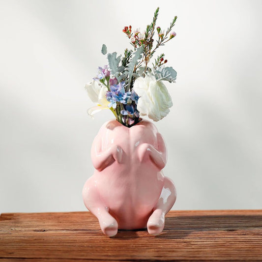 A pink ceramic raw chicken shaped vase filled with white, blue, and green artificial flowers, sitting on a rustic wooden surface against a soft white background.