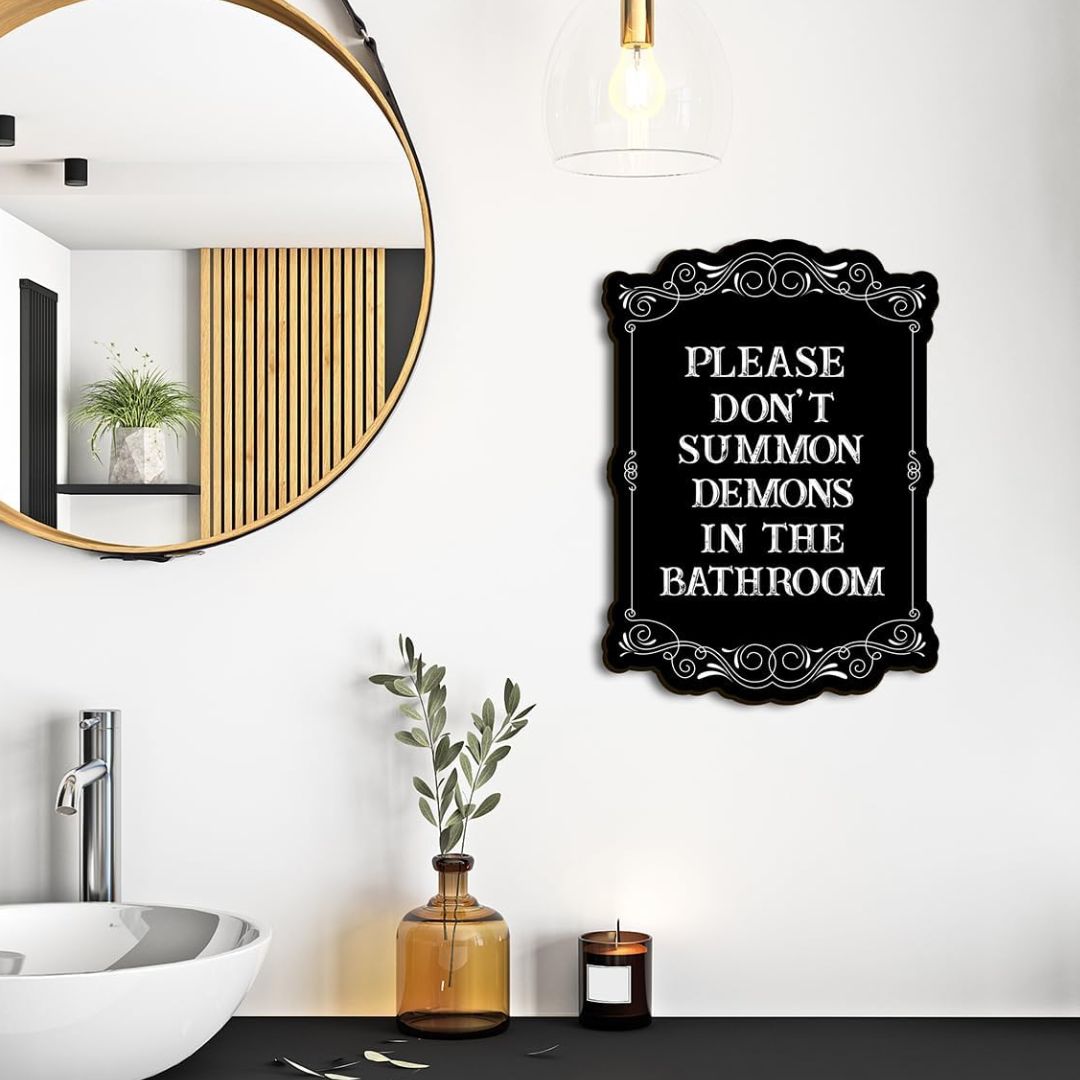 A black decorative wall sign with white text reading 'PLEASE DON'T SUMMON DEMONS IN THE BATHROOM' hanging on a white bathroom wall above a sink and countertop decor.