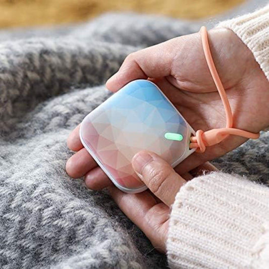 Hand holding a small, portable electronic hand warmer device with a geometric design on a textured surface.