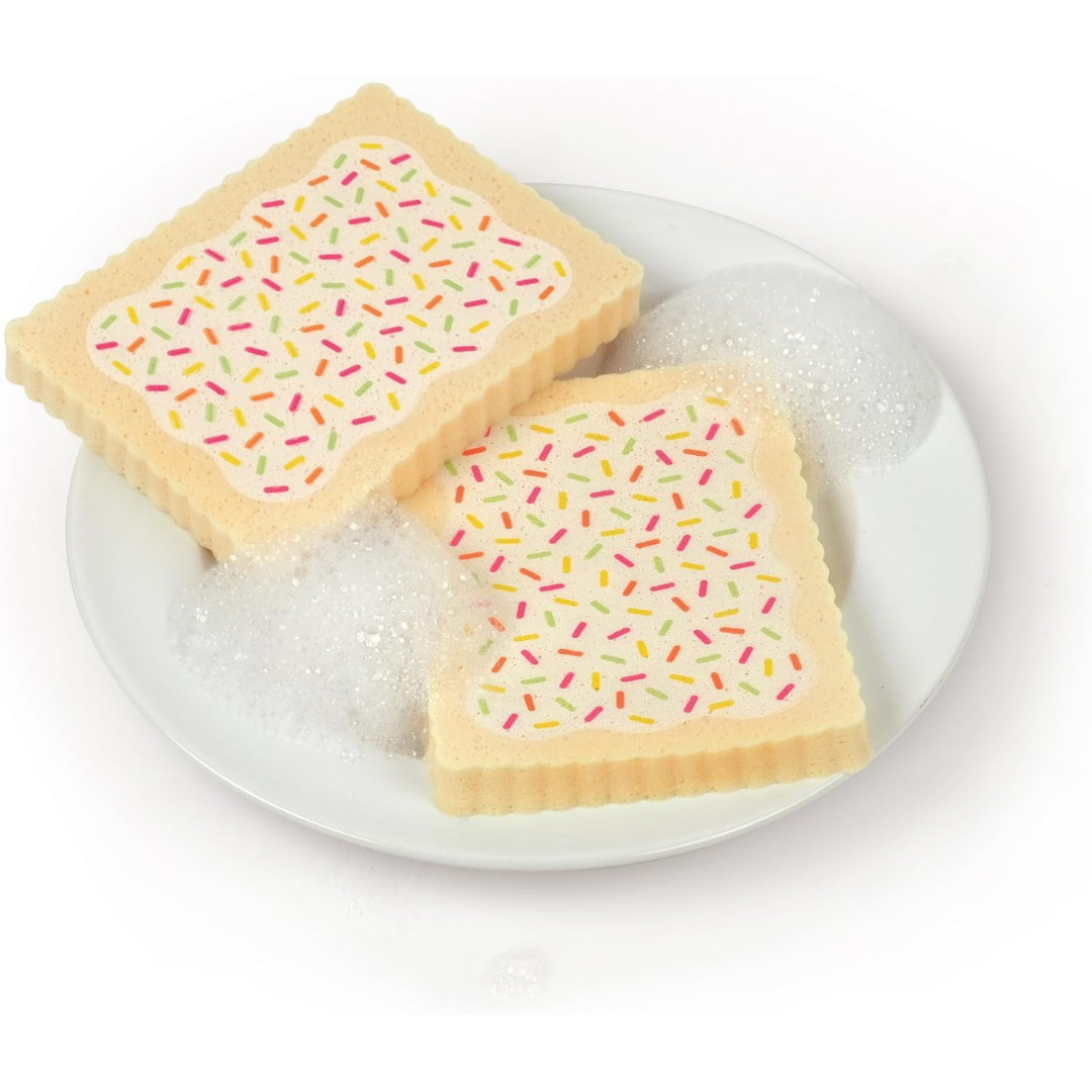 Two beige kitchen sponges shaped like Pop-Tarts with white icing and colorful sprinkles on a white plate, surrounded by soap bubbles, on a white background.