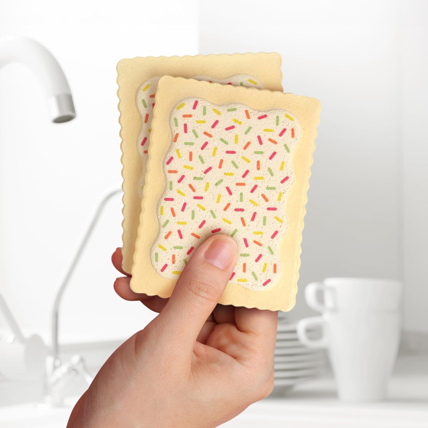 A hand holding two beige kitchen sponges shaped like Pop-Tarts with white icing and colorful sprinkles, in a kitchen with a sink and white dishes in the background.