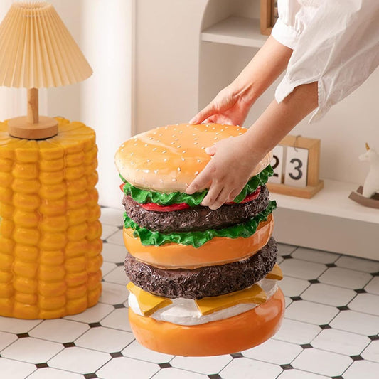 A person's hands are shown placing a hamburger shaped stool onto a tiled floor. The burger stool features a golden-brown sesame bun, two dark brown patties with green lettuce, red tomato slices, and yellow cheese.