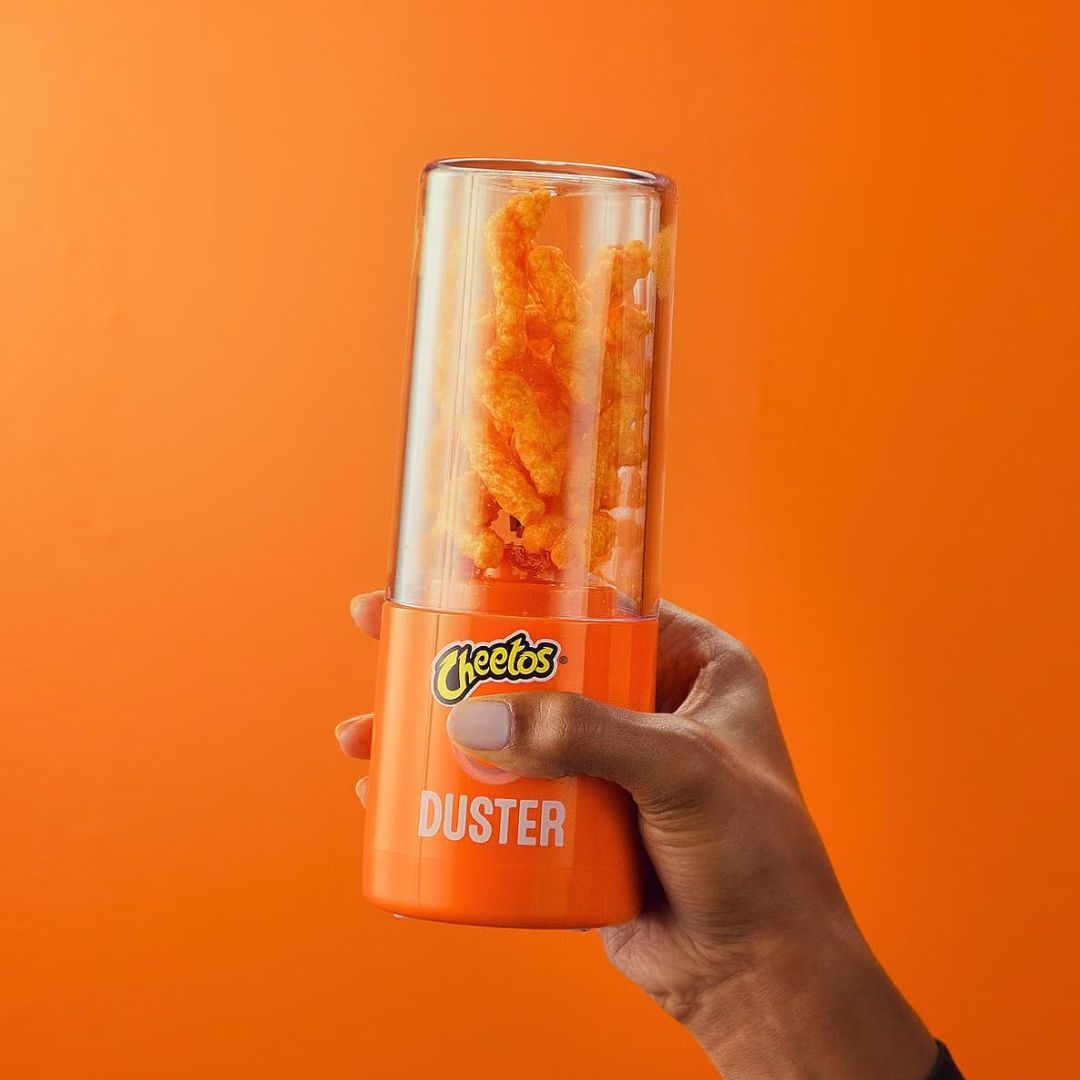 A hand holding a Cheetos Duster electric chip blaster against a solid orange background. The blaster has a bright orange base with the Cheetos logo and 'DUSTER' text, and a clear upper chamber containing orange Cheetos chips.