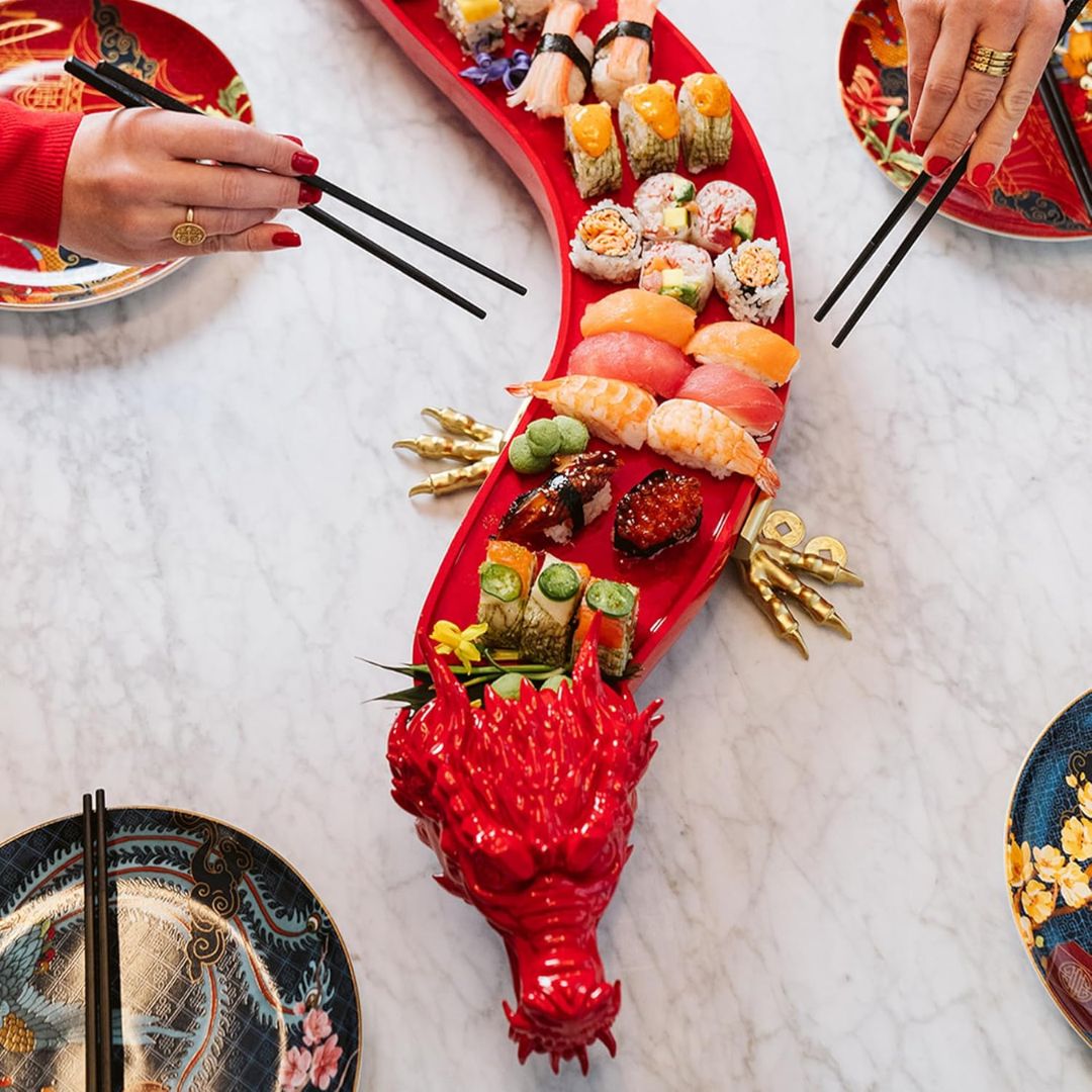 A red dragon-shaped sushi platter filled with assorted sushi rolls and nigiri, served on a white marble table with hands holding chopsticks around it.