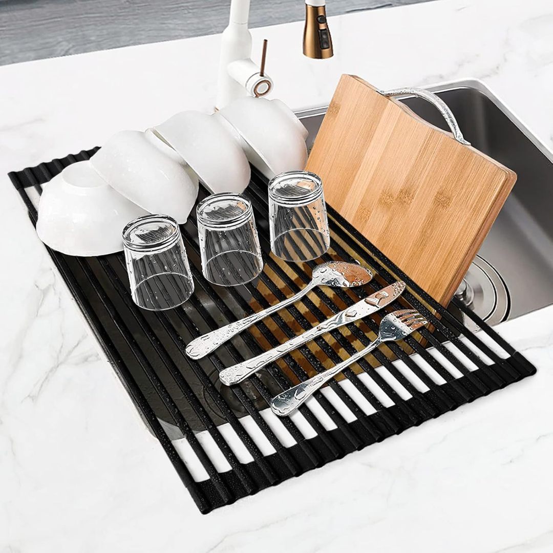Black roll-up drying rack over a kitchen sink holding bowls, glasses, cutlery and a wooden cutting board.