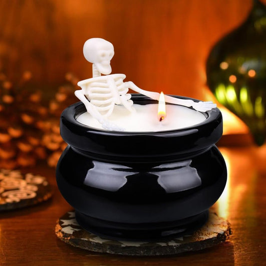 A black cauldron-shaped candle with a white skeleton figurine sitting inside, lit with a small flame, set on a wooden surface with warm holiday background lighting.