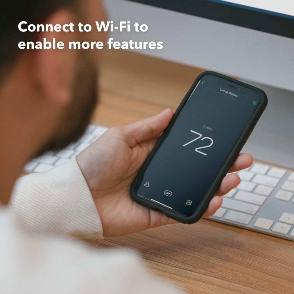 A hand holds a smartphone displaying an ecobee app with "Living Room" at 72°F and 40% humidity, on a desk with a keyboard. Text reads "Connect to Wi-Fi to enable more features."