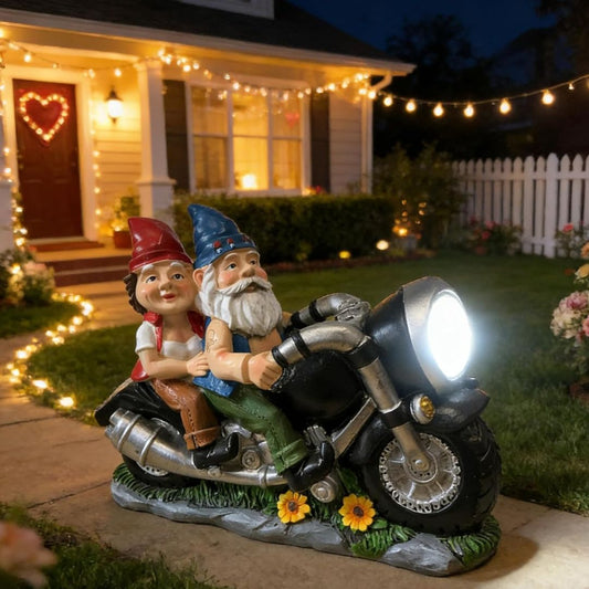 A decorative garden gnome motorcycle statue placed outdoors in a front yard at night. Two garden gnomes are sculpted on a motorcycle with a bright headlight, set on grass beside a house with warm string lights and a lit porch.