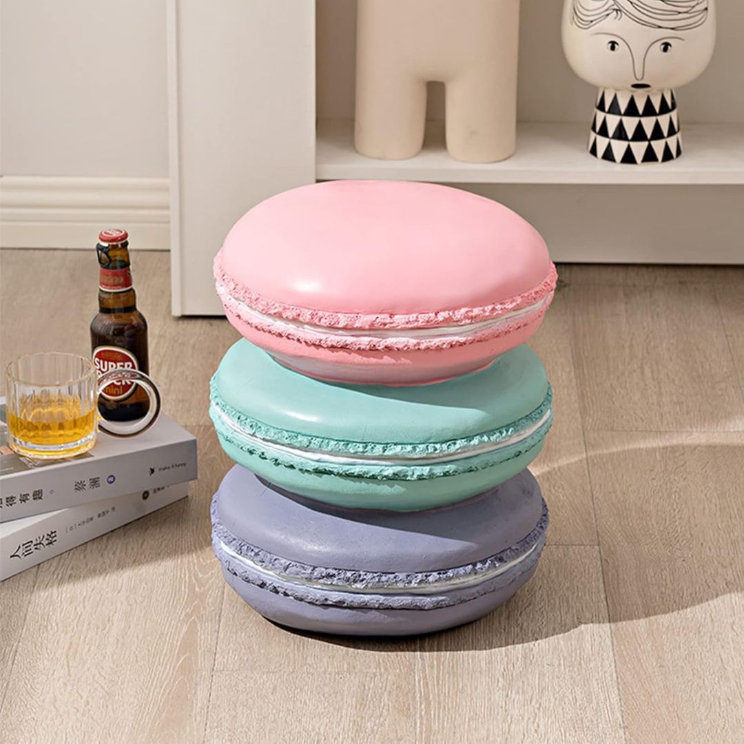 A stool shaped as three round macarons stacked vertically in pastel pink, green, and lavender on a light wood floor, with a glass of beer, books, and a shelf in the background.