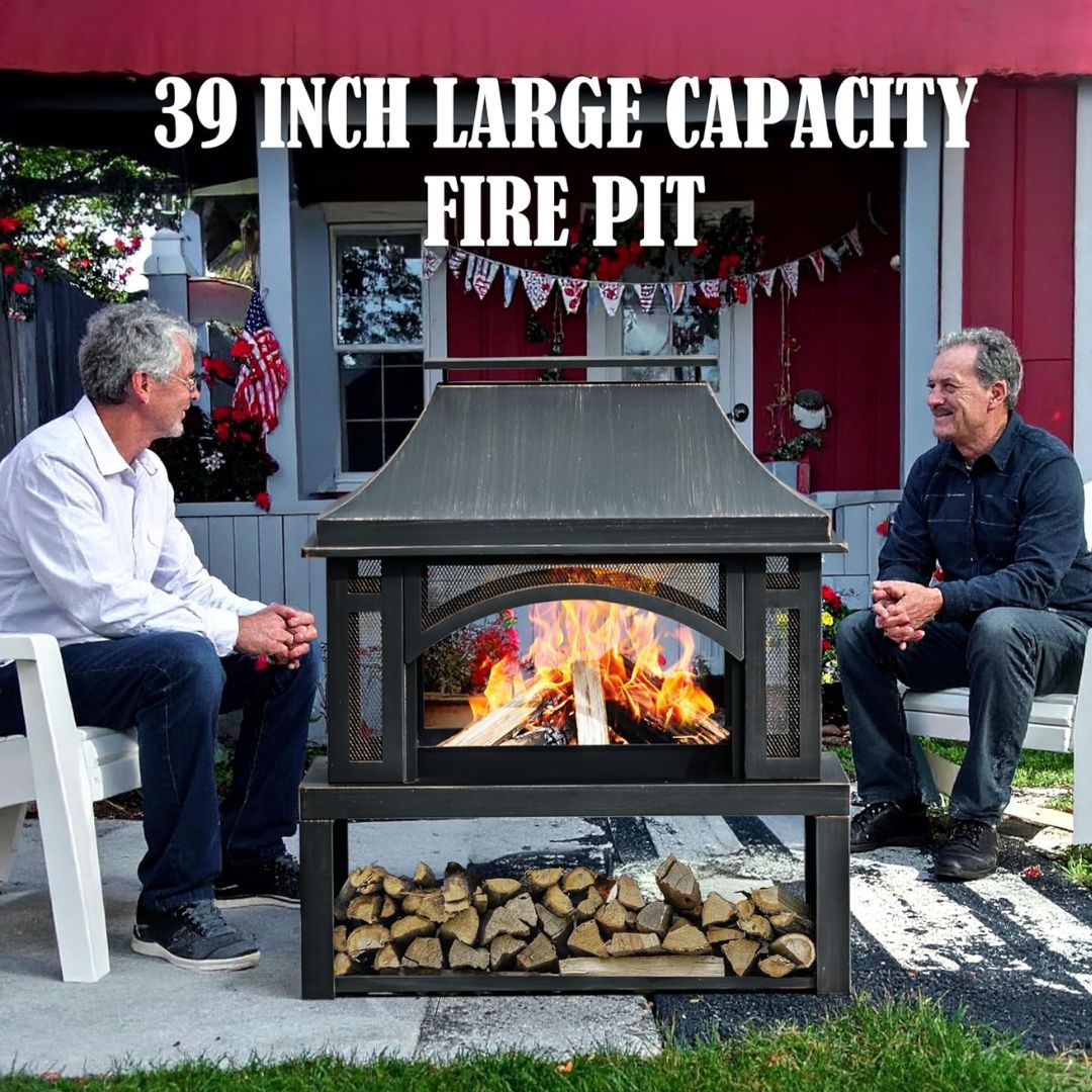 A large outdoor fireplace with an active fire inside, positioned between two men sitting on white chairs in a backyard setting. The fire pit has a dark gray metal frame, a slanted roof, a fire compartment with a glass front, and a lower shelf filled with stacked firewood.