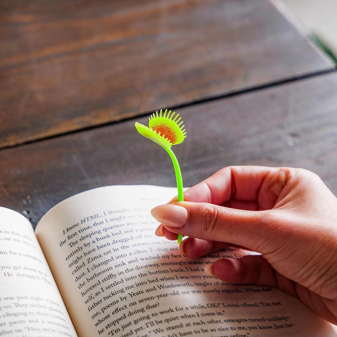 Person holding a green Venus flytrap bookmark over an open book on a wooden surface
