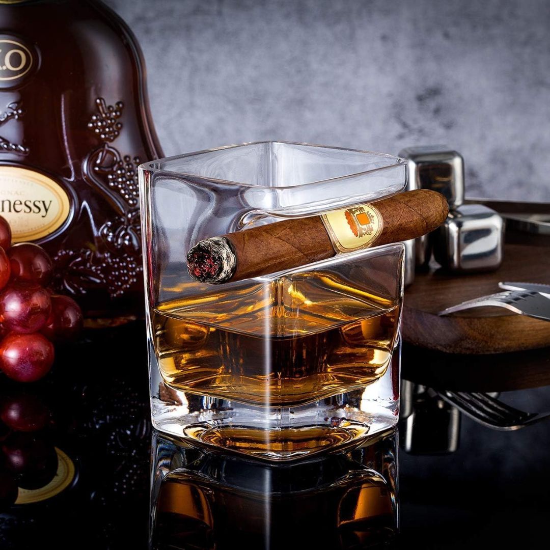 Whiskey glass with integrated cigar rest holding a lit cigar beside a bottle and bar accessories