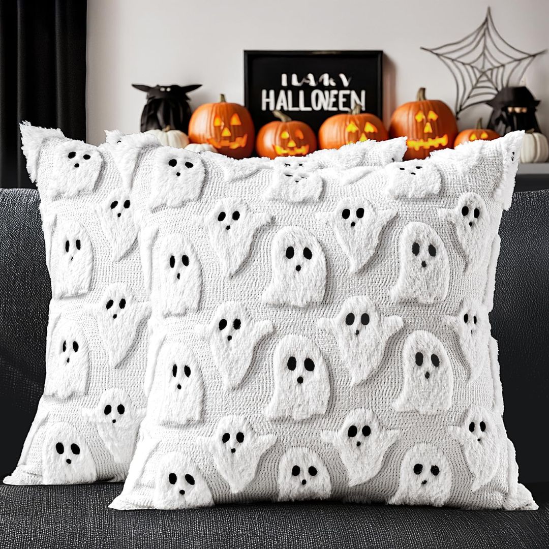Two white pillow covers with raised fluffy ghosts and black faces on a dark sofa, with Halloween pumpkins behind them.
