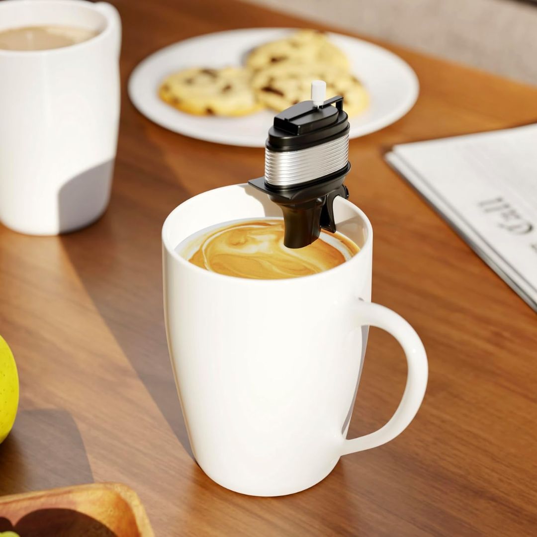 A white ceramic coffee mug on a wooden table, with a mini black and silver electric coffee boat motor stirrer inside the mug. A white plate with cookies and another cup are blurred in the background.