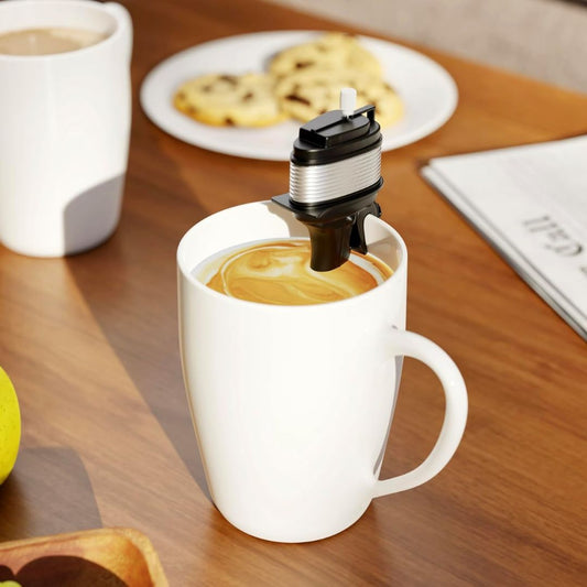 A white ceramic coffee mug on a wooden table, with a mini black and silver electric coffee boat motor stirrer inside the mug. A white plate with cookies and another cup are blurred in the background.