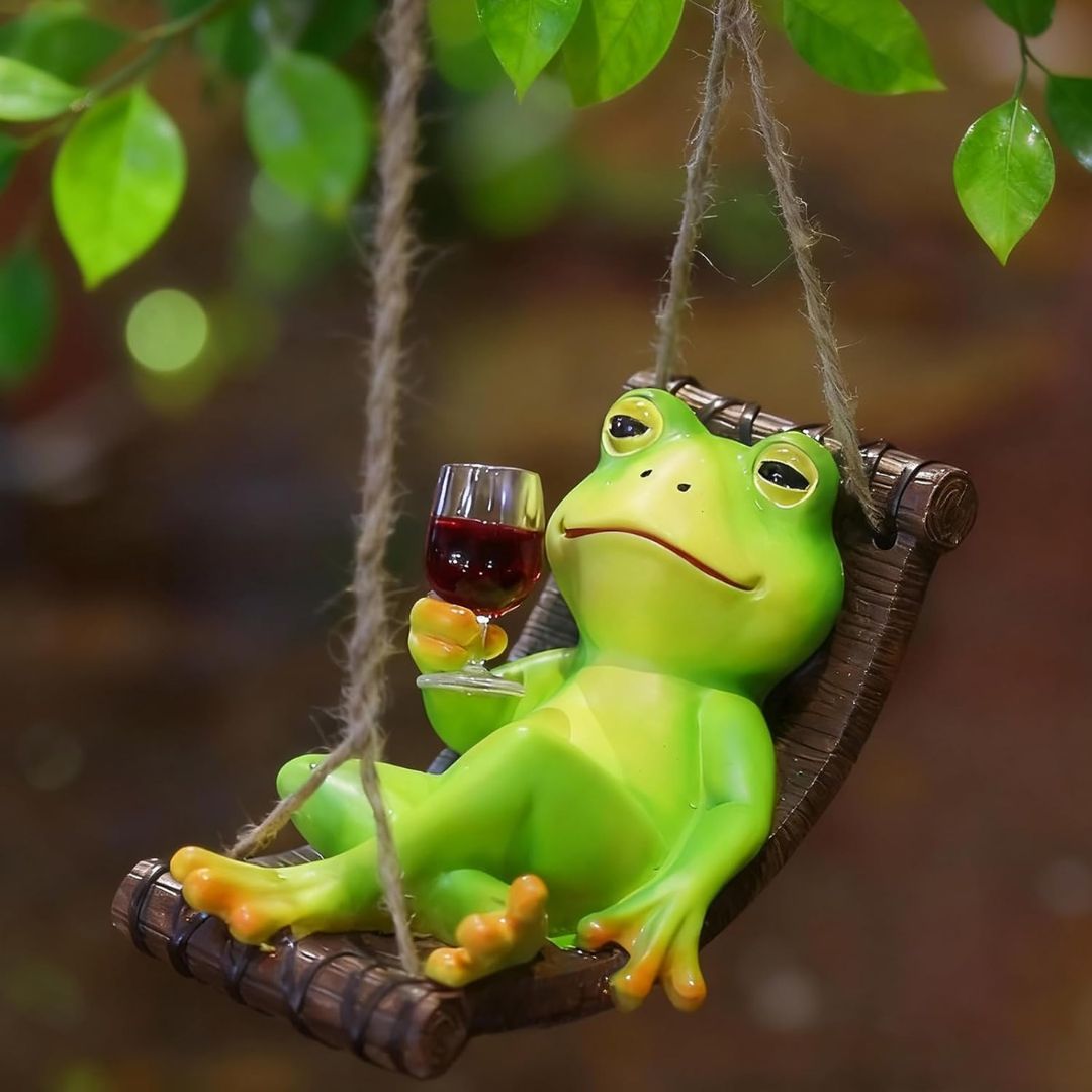 A green frog figurine reclining in a brown hammock swing and holding a glass of red wine, set against a blurred background of green leaves and brown bokeh.