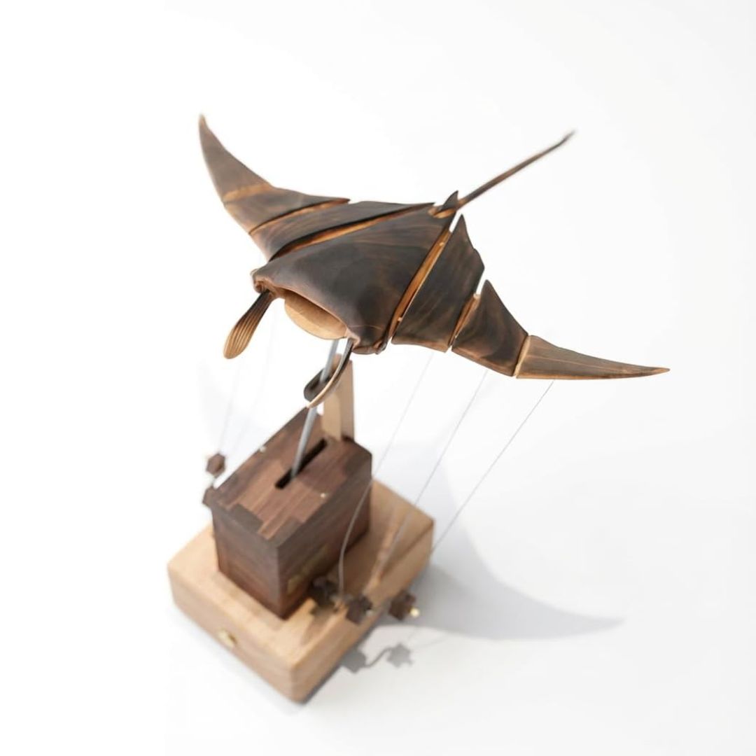 A wooden manta ray sculpture mounted on a small wooden base with string and pulley details, photographed on a clean white background.