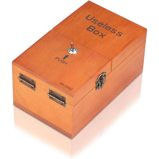 A wooden 'Useless Box' with a warm orange-brown finish, featuring a silver metal switch on top, latches on the sides, and printed text on the lid and front.