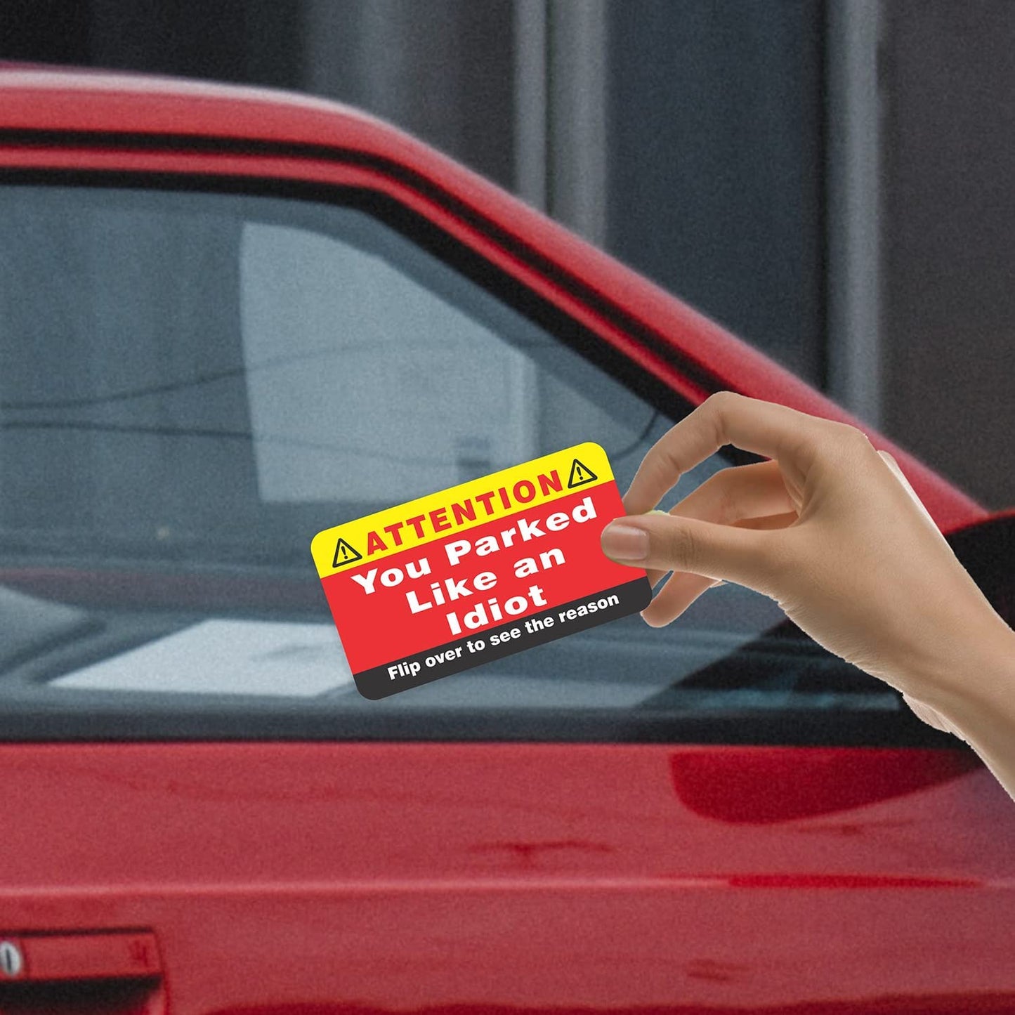 A hand is holding a business card in front of a red car. On the front of the card it reads, 'You parked like an idiot, flip over to see the reason.'
