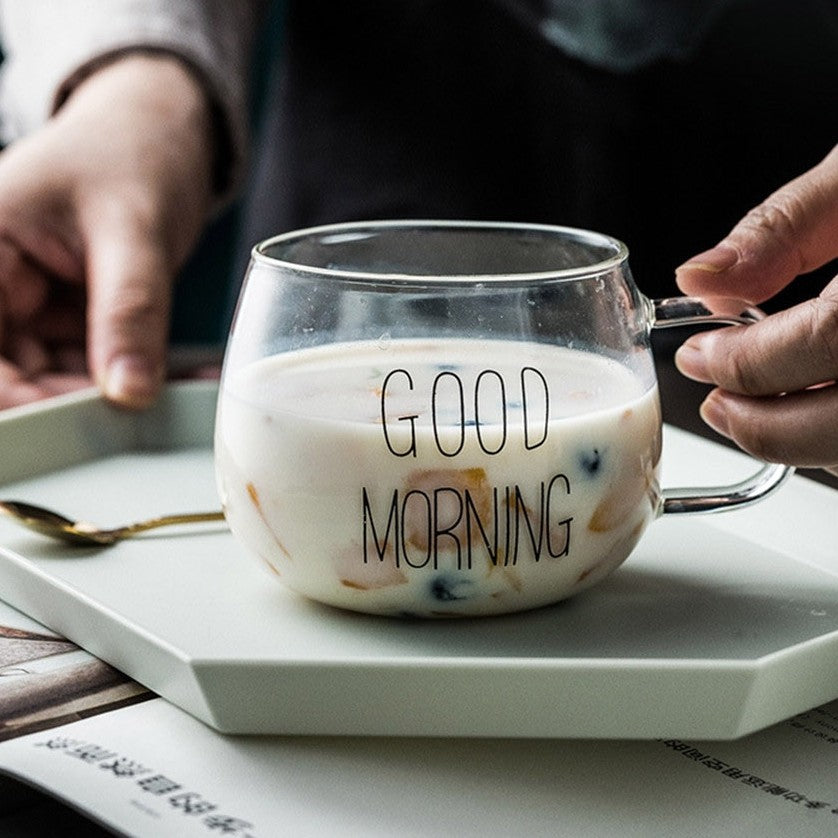 A hand about to pick up a good morning glass mug with milk and cereal off a ceramic plate