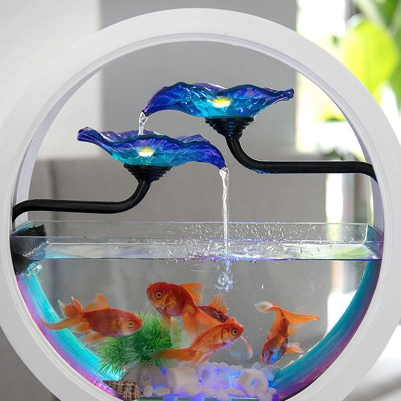 A close-up view of a waterfall fish tank air purifier. The fish tank is white and circular. Inside are five goldfish. Water is trickling down two blue glass flowers and back into the aquarium. There is a blue and purple light inside the aquarium with a sea shell and fake green plant also inside the tank.